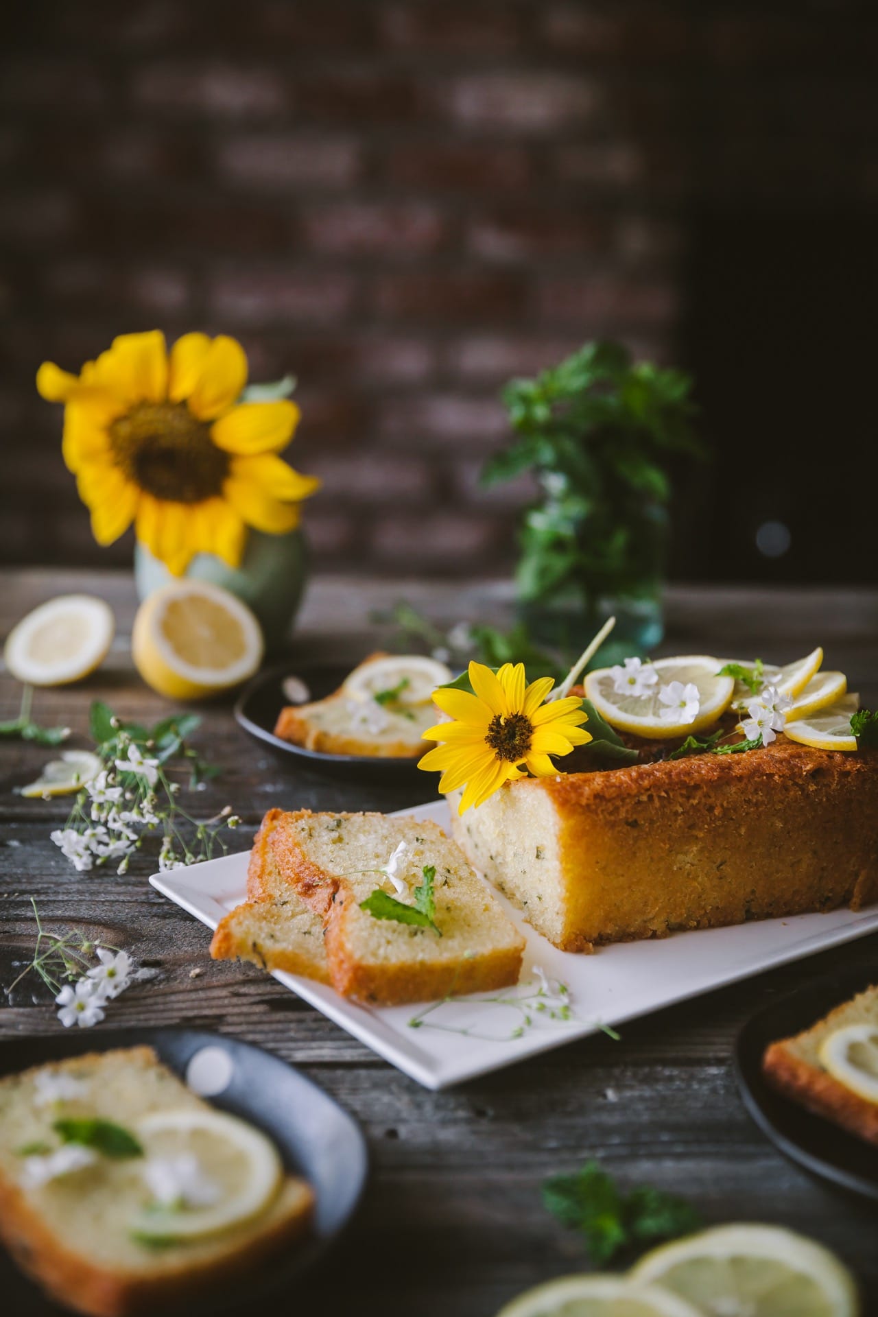 Lemon Mint Cake styled with flowers and fresh lemon and mint. It's sliced and the slices are kept next to the rest of the cake loaf