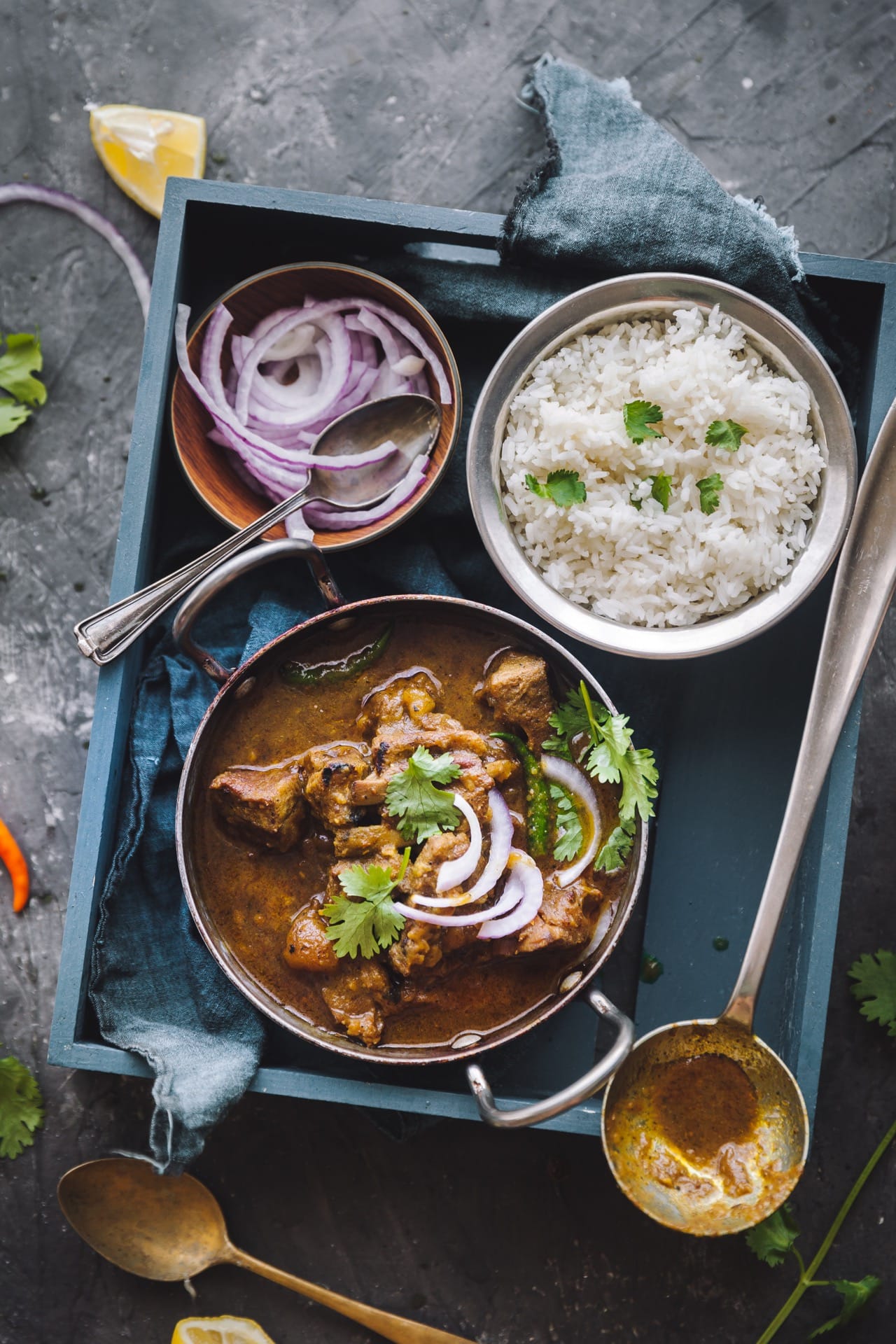 Over the top view of Sunday Mutton Curry in a bowl served with rice in a different bowl along with sliced onion. Everything styled on a blue wooden tray