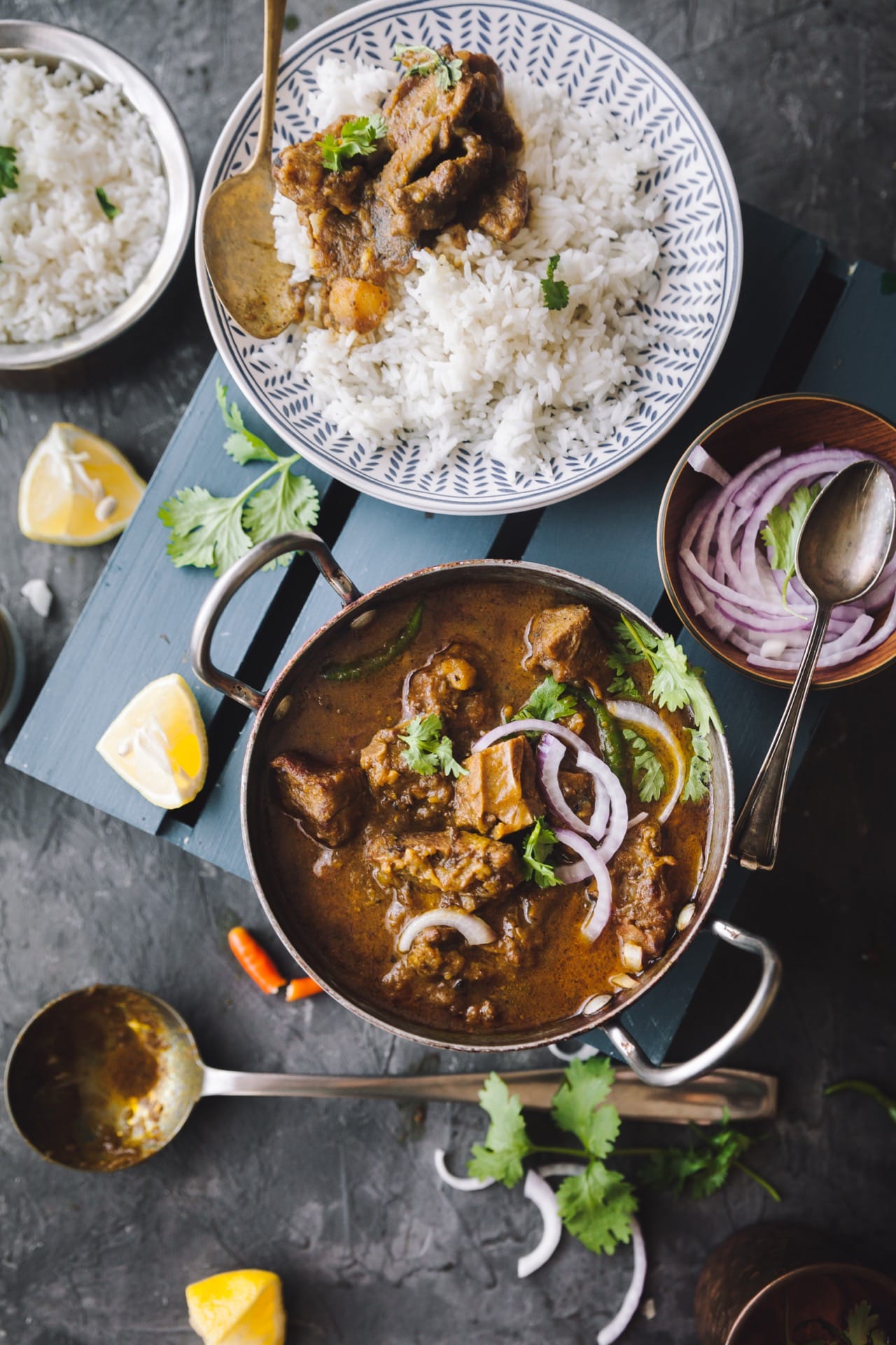 Over the top view of Sunday Mutton Curry along with a plate with rice and curry on the side. Sliced onion in a bowl and rice in a bigger bowl.